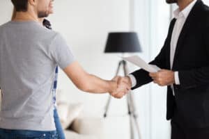 Two people are shaking hands indoors. One is wearing a suit and holding papers while the other is dressed casually in a grey t-shirt and jeans.