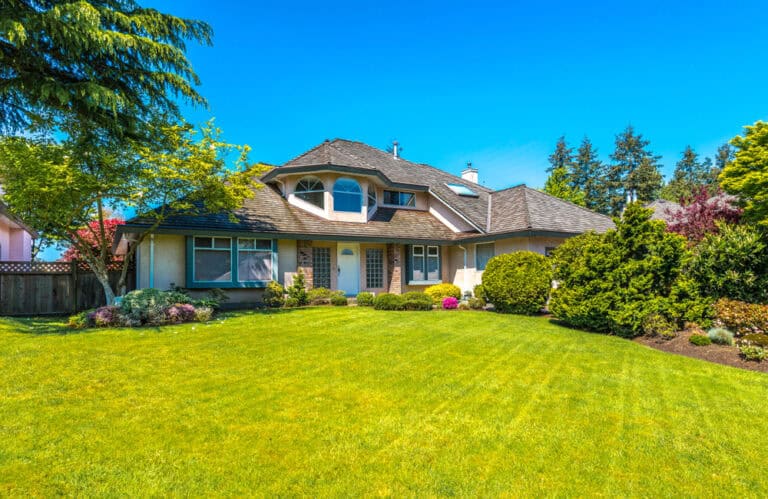 A charming two-story house with a well-maintained lawn, surrounded by lush greenery and trees, under a clear blue sky.