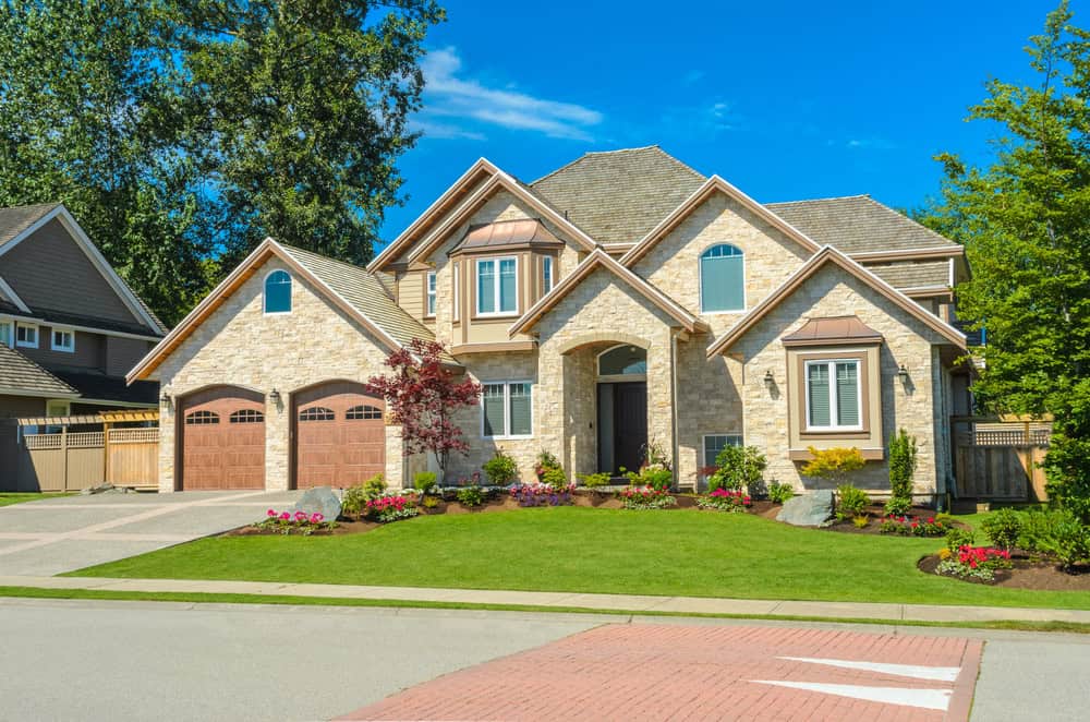 A modern, two-story stone house with a neatly landscaped front yard, double garage, blue sky, trees, and greenery surrounding the property.