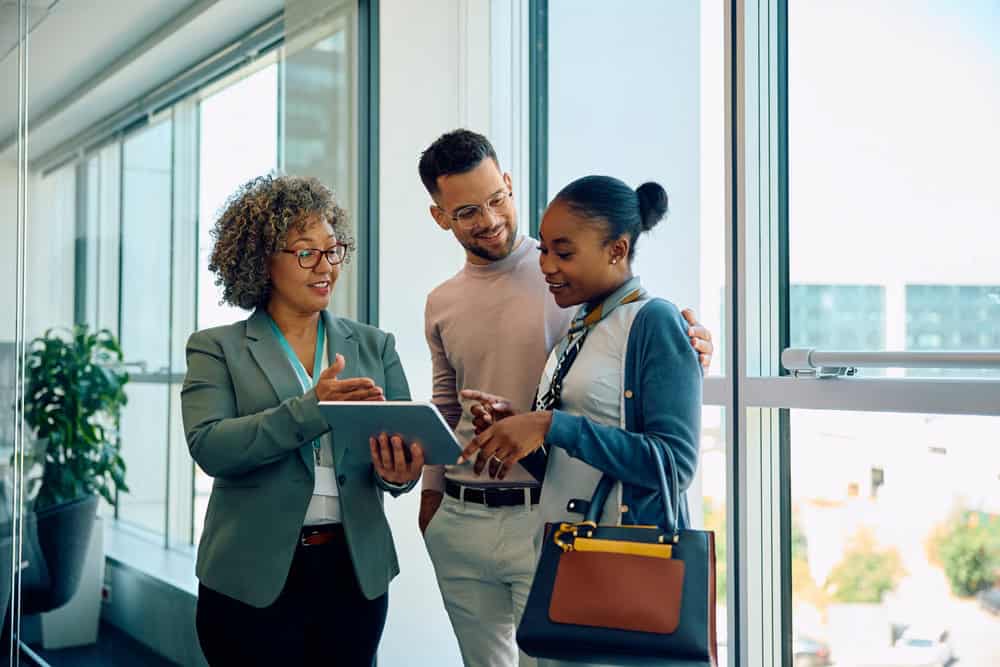 Three professionals standing and discussing a document on a tablet near a window in an office. One woman holds the tablet while others listen attentively.