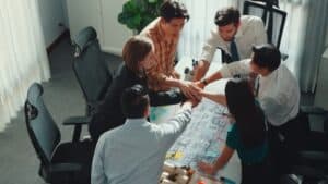 Six colleagues standing and putting their hands together over a table with blueprints and notes, signifying teamwork in a well-lit office.