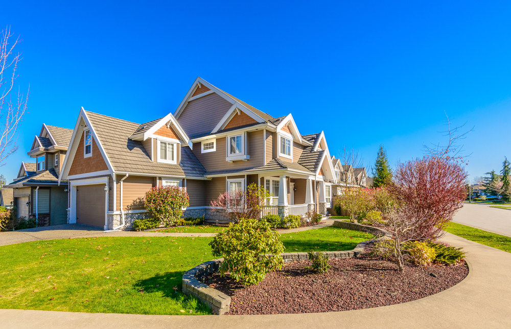 A large, two-story house with a well-manicured lawn, shrubs, and trees. The sky is clear and blue on a sunny day. A driveway is on the left.