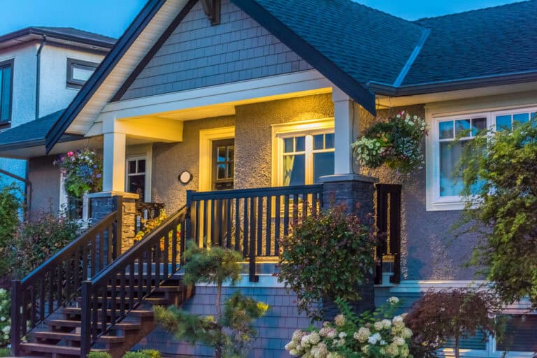 A cozy house at dusk with a lit front porch, dark railings, and steps leading up; flowers and greenery adorn the entrance.