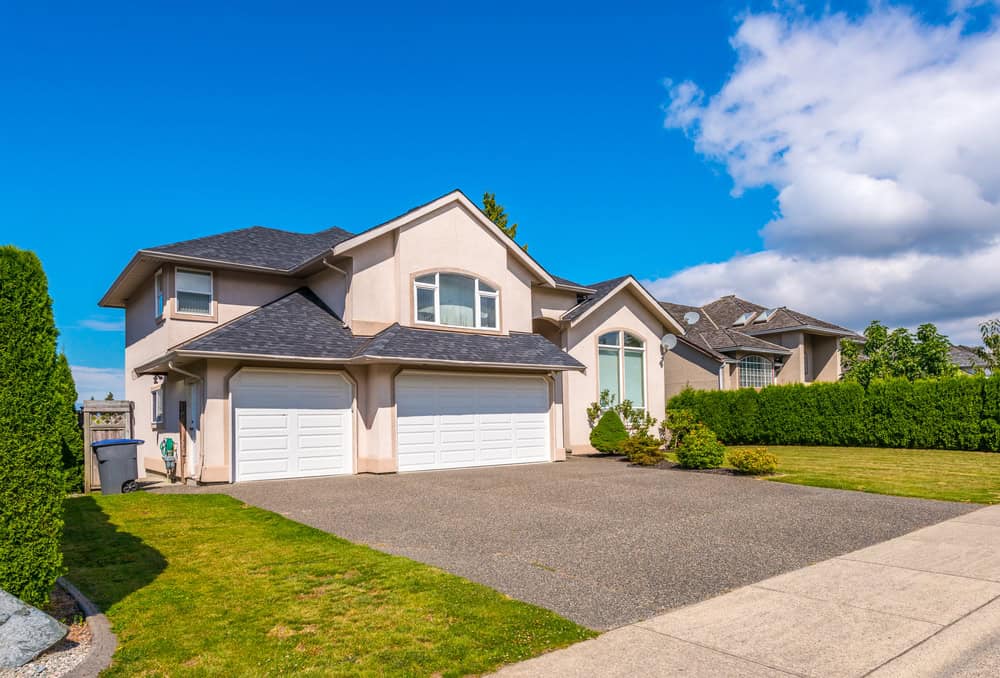 A beige two-story house with a double garage, green lawn, and blue sky with a few clouds. Adjacent houses and hedges are visible.